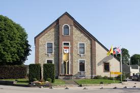Scène urbaine à Manhay avec entreprise de débouchage, rue calme bordée d'arbres et maisons en pierre typiques de la région